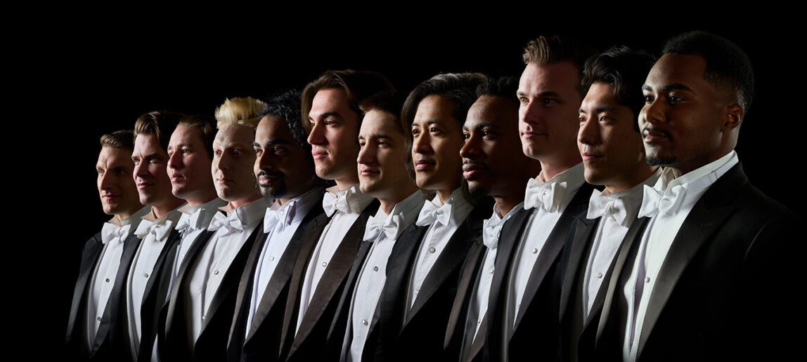 Twelve members of a vocal ensemble, all wearing tuxedoes, are lined up next to each other.