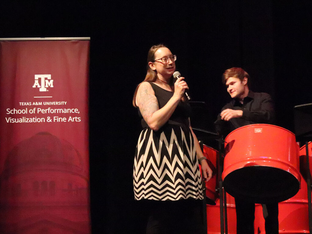 A university professor speaks into a microphone onstage. To her left is a student steel-plan player. To her right is a maroon banner that reads: Texas A&M University, School of Performance, Visualization & Fine Arts.