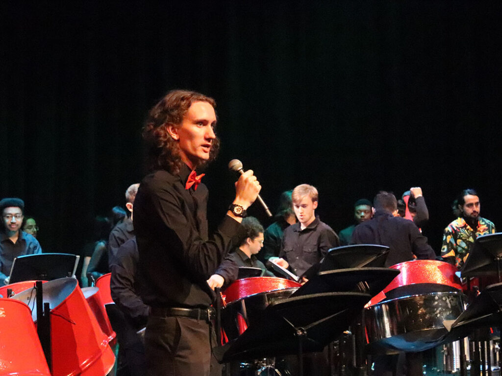 A college student wearing black and a red bow tie speaks into a microphone onstage. Behind him is a steel-pan ensemble.