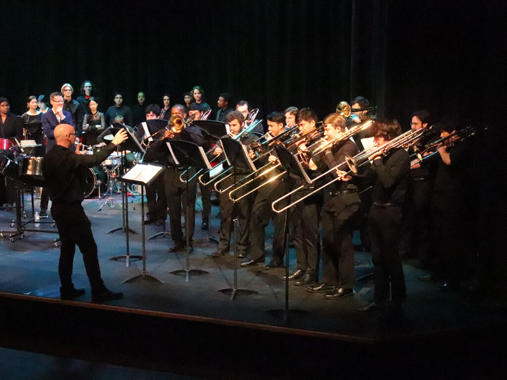 Trombone players stand on a stage, performing alongside steel-pan ensemble students. A conductor stands before them.