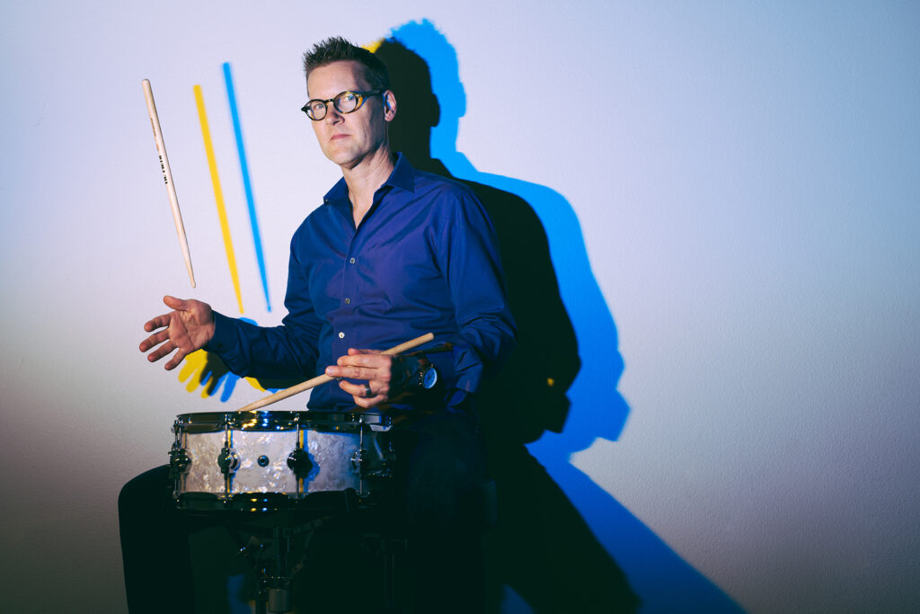 A percussionist is shown playing a drum, with one drumstick in the air.