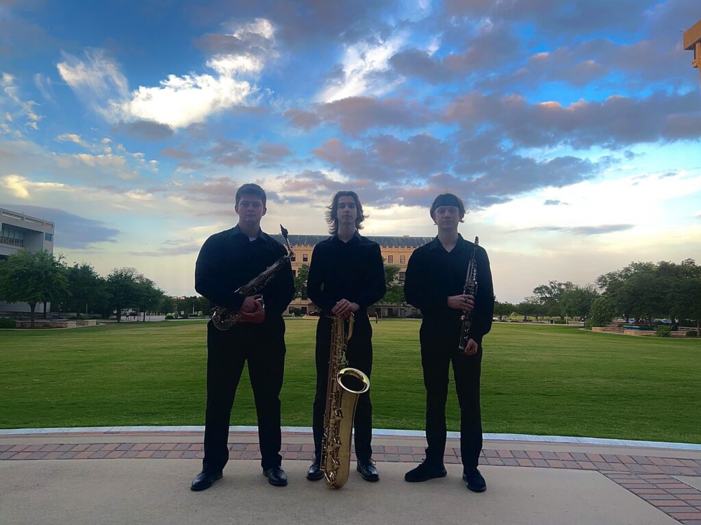 Three college students hold their instruments as they stand before a large field.