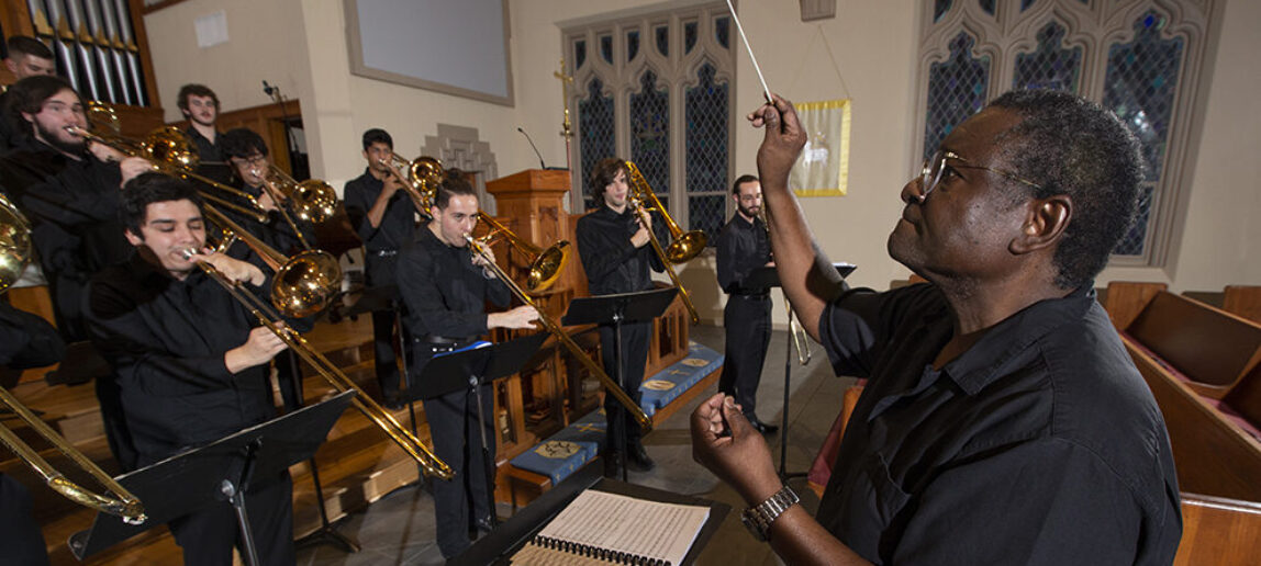 A music professor standing on the right conducts a choir of trombone players.