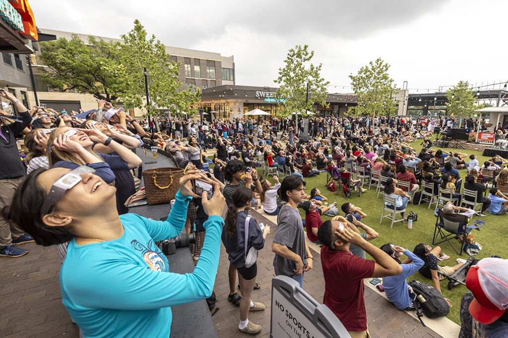 A large crowd of visitors wearing solar glasses looks up during the solar eclipse.