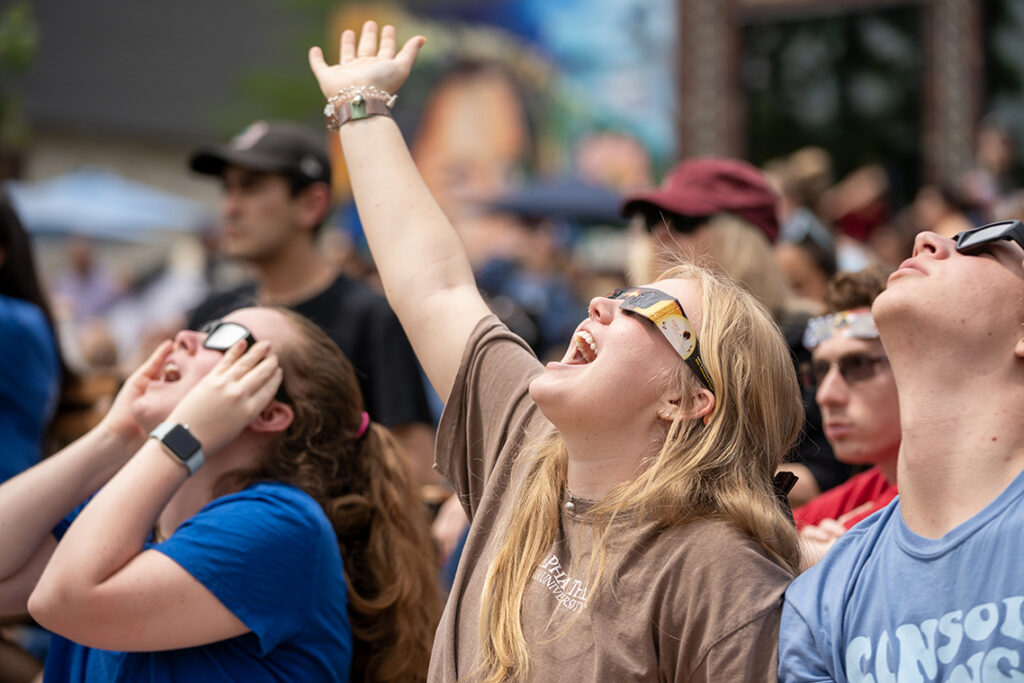 A student wearing solar glasses smiles and holds her right arm up toward the sky during the solar eclipse.