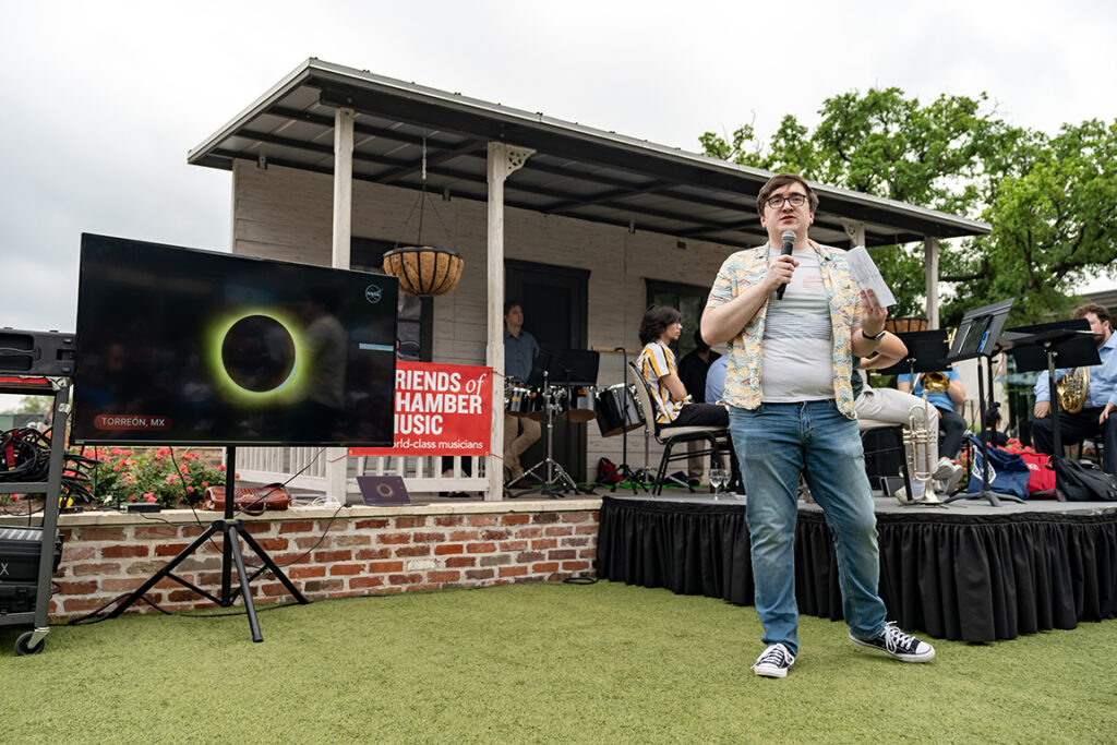 A college professor speaks by a stage during an event for the solar eclipse. To his right is a TV screen showing an eclipse scene.