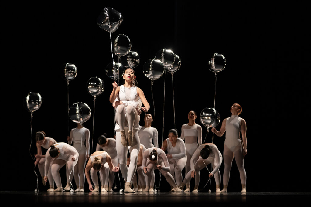 Two members of a high school dance troupe are shown, with one picking the other up into the air. Behind them are multiple members of the troupe. All are wearing white, and holding silver balloons.