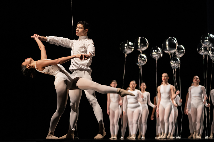 Two members of a high school dance troupe, both wearing white, are shown performing onstage. In the background are multiple members of the troupe, all wearing white and holding silver balloons.