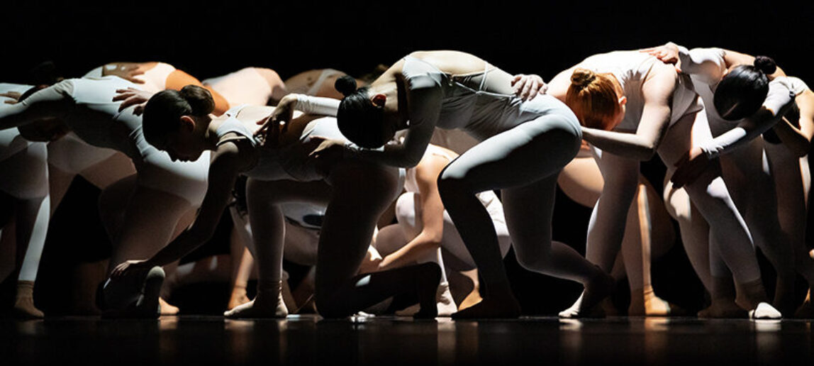 Members of a high school dance troupe, all wearing white, are shown performing onstage. They are all crouched down toward the floor, with their arms on the dancer in front of them.