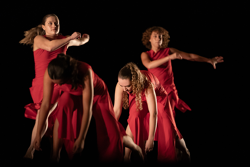 Four members of a high school dance troupe, all wearing red, are shown performing onstage. Two are bent over toward the floor, and two are standing behind them.