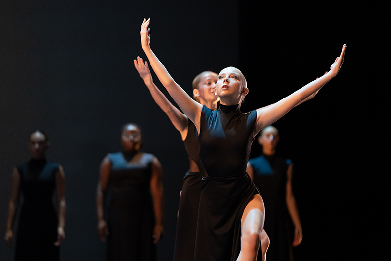 Two members of a high school dance troupe are shown performing onstage, with their arms stretched up. Three members are shown standing in the background.