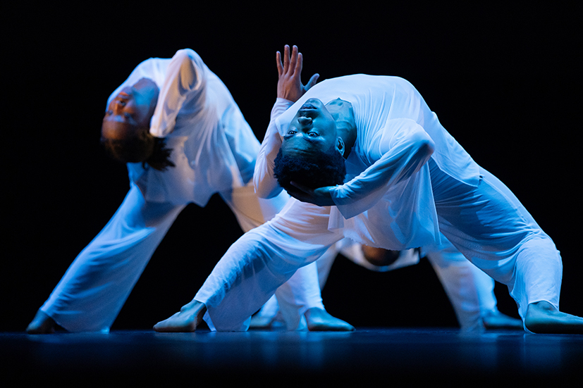 Three members of a high school dance troupe perform, all wearing white. They are bent over backward, with their faces to the camera.