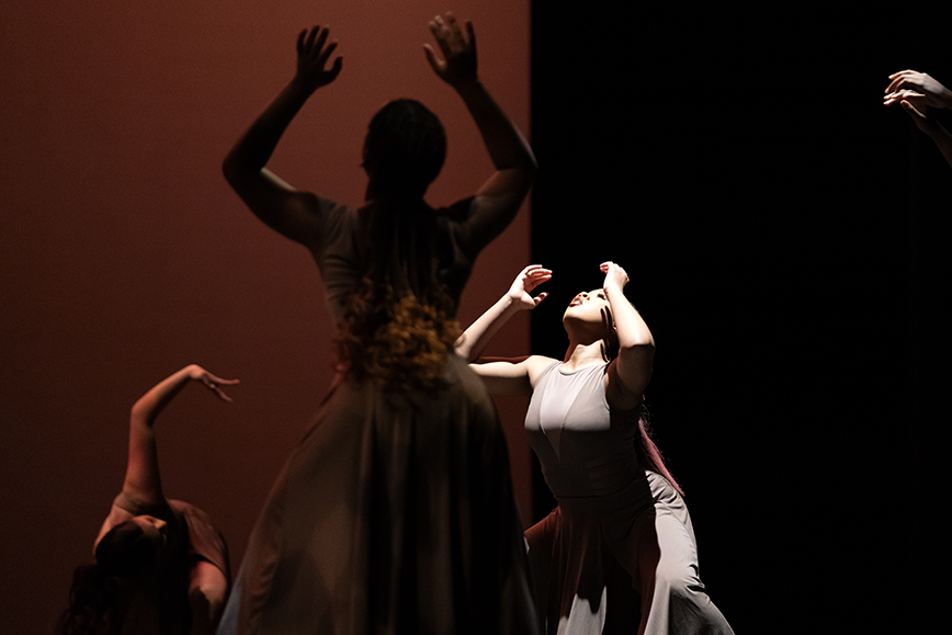 Four members of a high school dance troupe are pictured, reaching toward their heads and looking up.