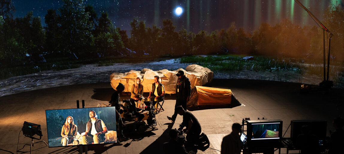 Two actors are on a virtual production stage shooting a movie. The camera view is visible on a screen to the left. The images behind them make it appear they are in a nighttime wilderness scene. Crew members are shown working in the background.