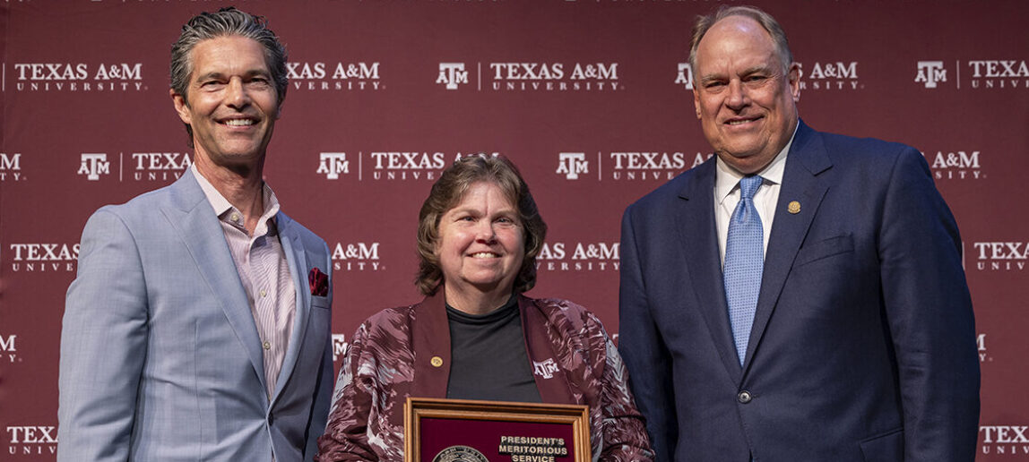 A university employee holds a President's Meritorious Service Award with two university leaders standing on either side of her. A maroon backdrop with Texas A&M University logos is behind them.