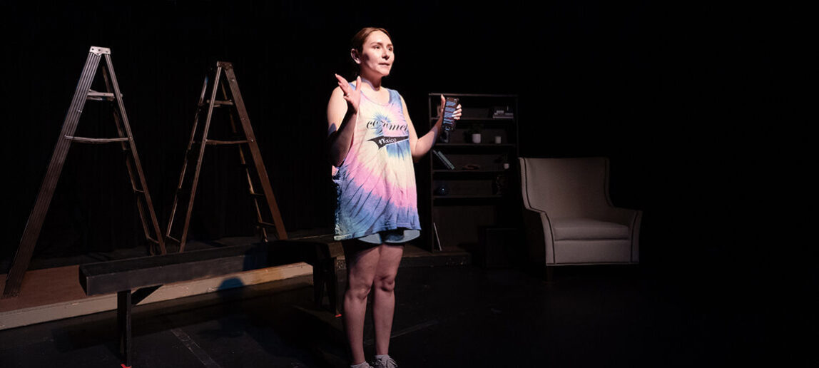 A college student who serves as the director of a musical stands on a stage as she addresses her cast and crew.