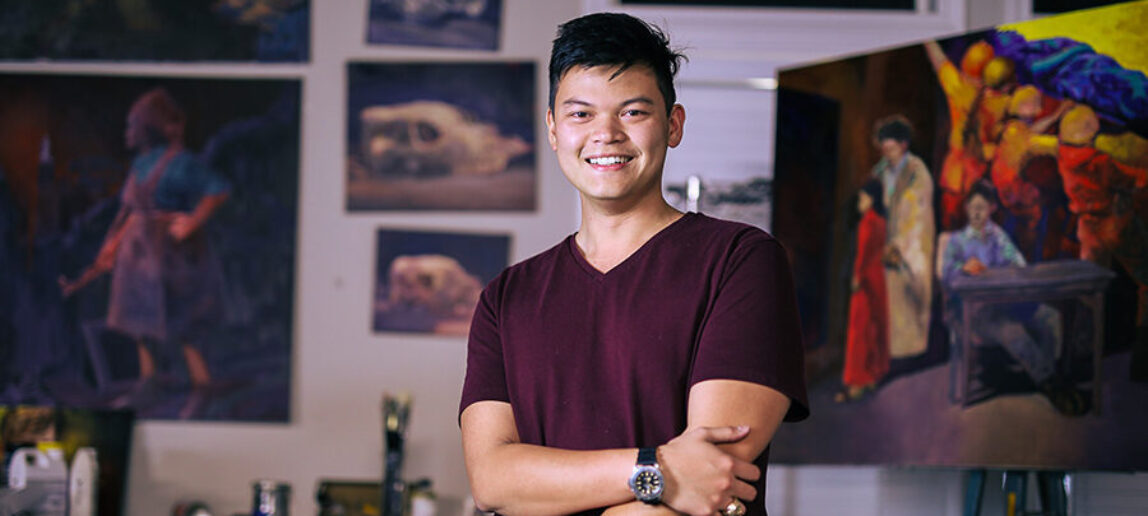 A college student stands in front of a wall full of of paintings.