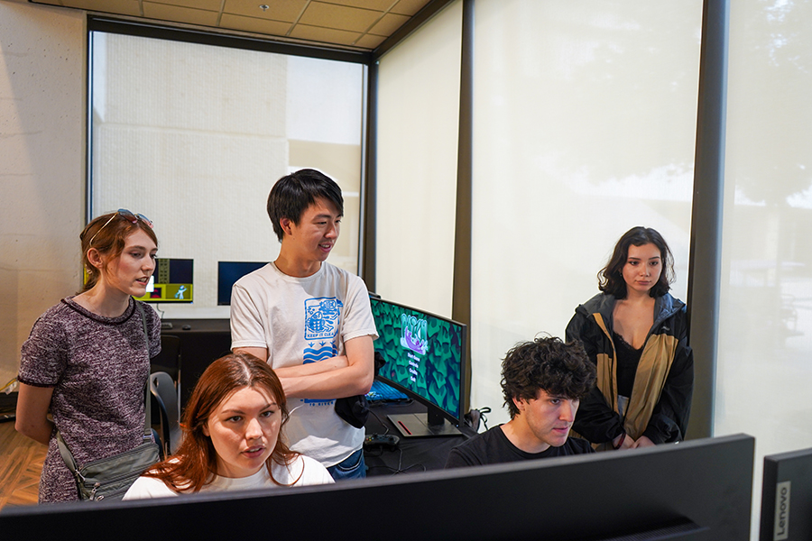 Two college students play video games, while three students stand behind them to watch.