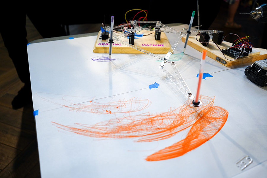 Colored pens that are connected to a device labeled "Drawing Machine" make colorful designs on a large piece of paper.