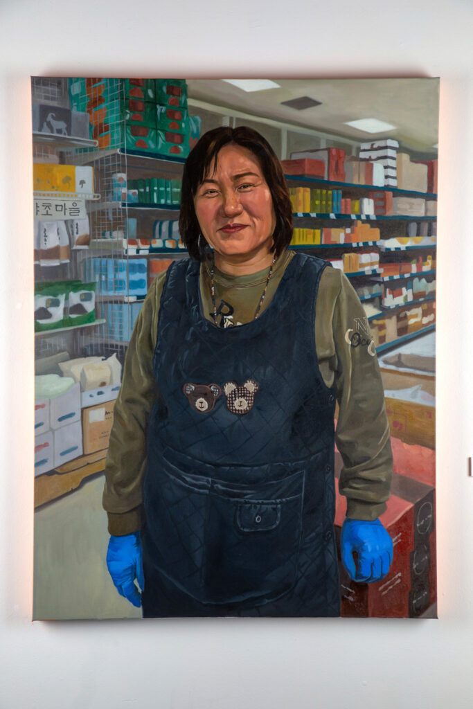 A painting that depicts a Korean woman wearing overalls and work gloves in a grocery store.