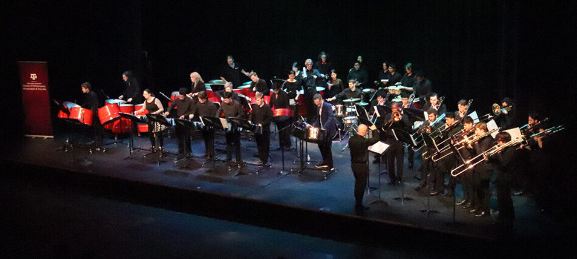 A group of musicians perform on a stage. The performers on the left are playing steel pan, and the performers on the right are playing trombone.