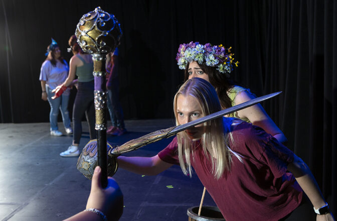 A college student onstage holds a sword close to her face, while a student behind her with a flower head band looks worried.