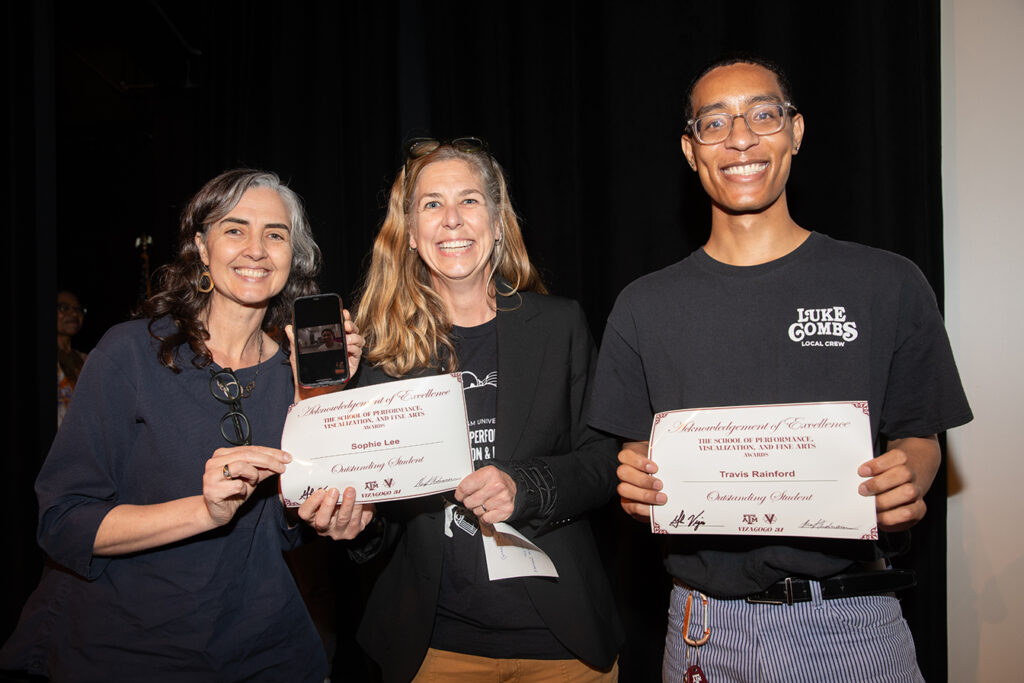 Two college professors stand by a student holding an award certificate. The professor on the left is also holding a phone with an award winner on Facetime.