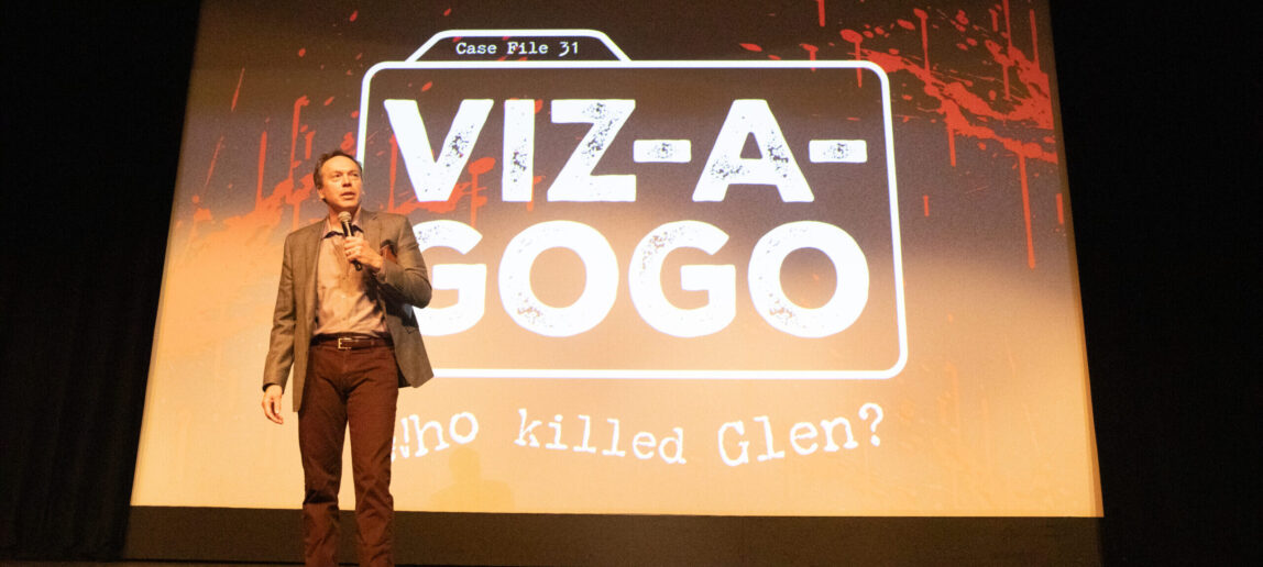 A college dean stands on a stage, speaking into a microphone. Projected behind him on a screen is a graphic that says Case File 31. Viz-A-GoGo. Who Killed Glen?