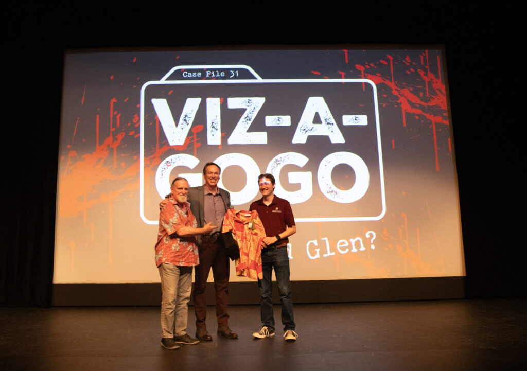 Three people are on a stage, all holding up a Hawaiian shirt. Projected behind them on a screen is a graphic that says Case File 31. Viz-A-GoGo. Who Killed Glen?