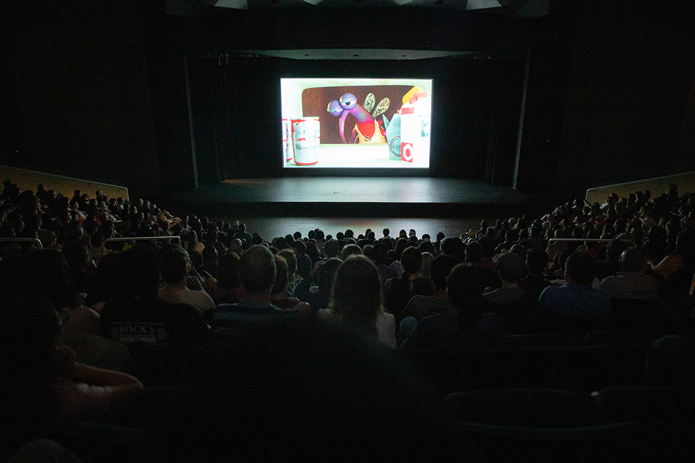 A crowd of people in a theater watch an animated short featuring a mosquito character.