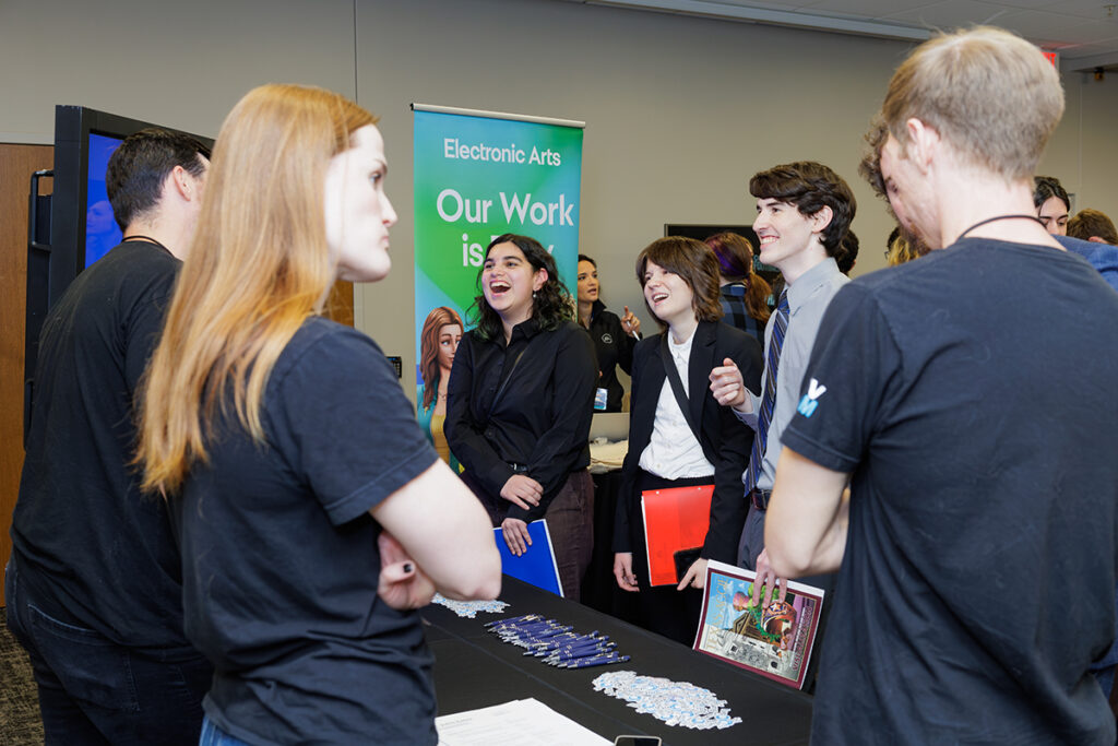 A group of college students are gathered at a table at a job fair, talking to industry representatives.