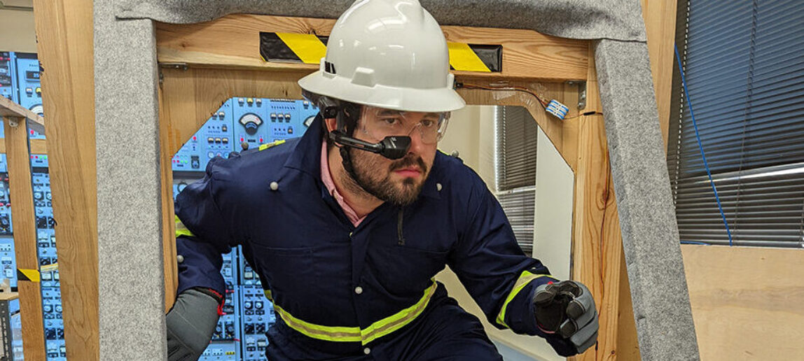 A professor dressed as a maritime surveyor wears a hard hat and augmented reality headset device as he steps through a simulated environment.