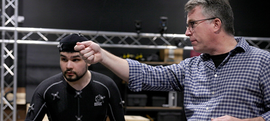 A college student wearing a black suit outfitted with sensors works with a professor at a motion capture facility.