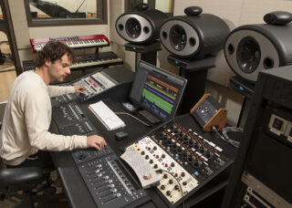 A student sits at a console working with a sound board and other music equipment.