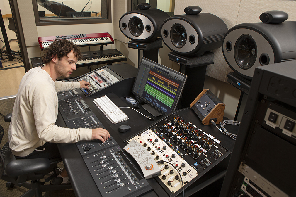 A student sits at a console working with a sound board and other music equipment.