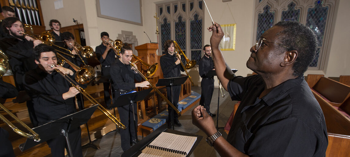 A music professor conducts an orchestra of trombone players in a church setting.