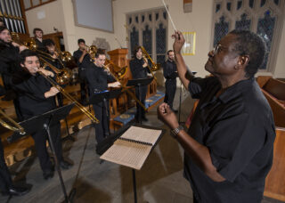 A music professor conducts an orchestra of trombone players in a church setting.