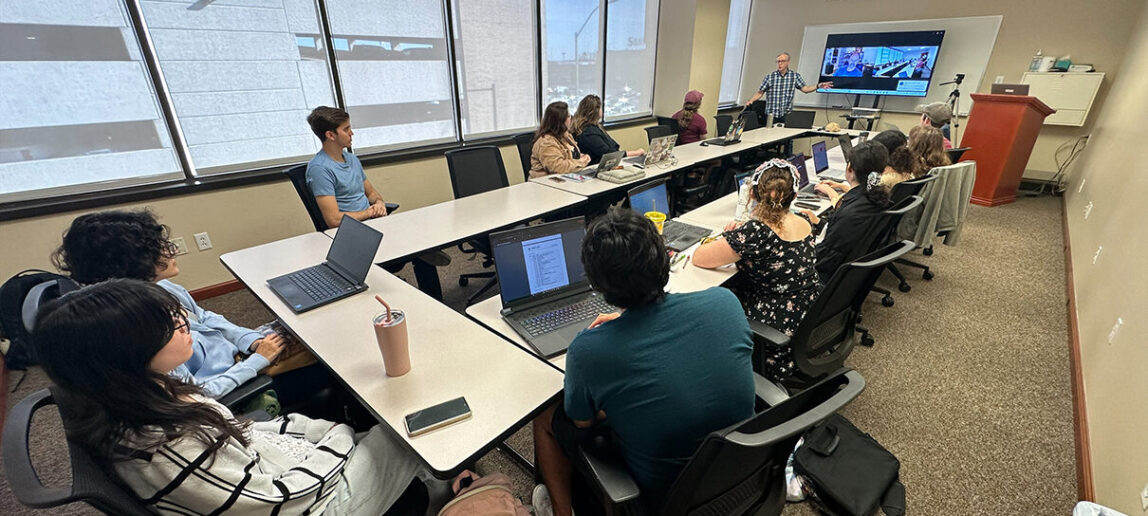 College students are seated around a long rectangular arrangement of tables next to a wall of windows. The students are listening to a lecturer speaking at the end of the table. Behind the lecturer is a screen showing another person joining the conversation via Zoom.