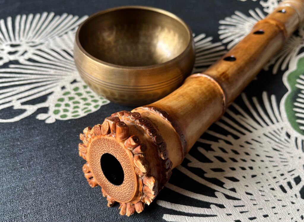 A shakuhachi, an end-blown bamboo flute, is shown on a table with a decorative design and a gold bowl.