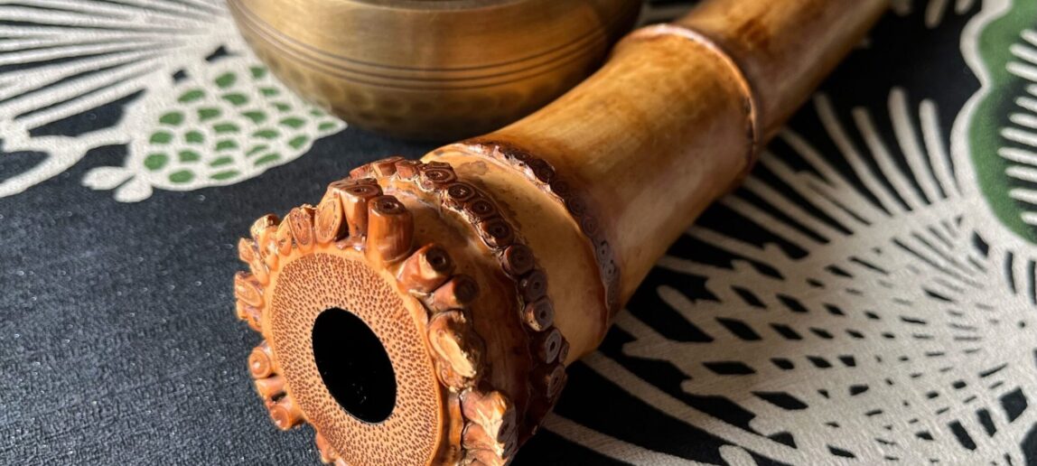 A shakuhachi, an end-blown bamboo flute, is shown on a table with a decorative design and a gold bowl.