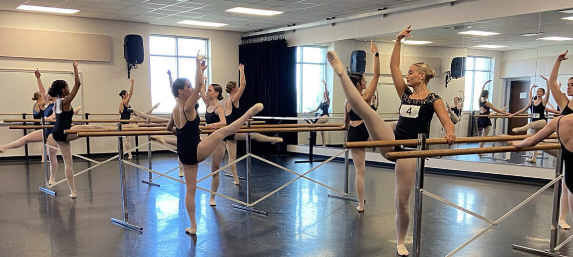 High school dance students practice their movements as they stand by the barre in a dance studio.