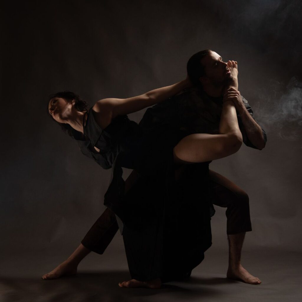 Two dancers perform in a dark studio environment.