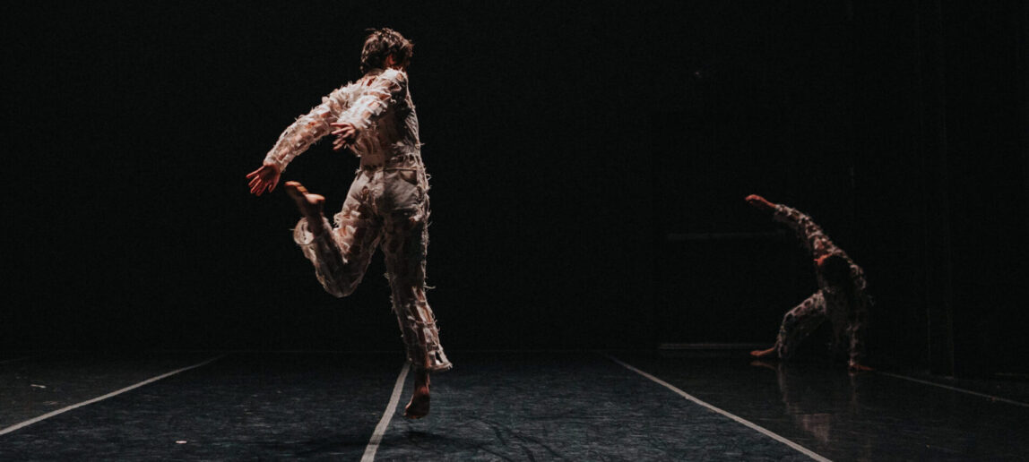 A dancer is shown from the back in a darkened stage environment. The dancer is leaping off their right foot, with their left leg bent behind them. To the right another dancer performs, balancing on their left arm and leg.