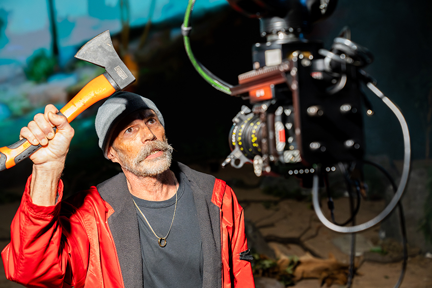 An actor wearing outdoor camping gear holds up an ax with his right hand as he looks into a video camera on a virtual production stage.