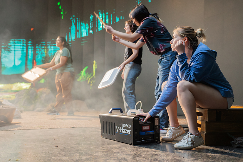 College students hold props for creating effects for a short film created on a virtual production stage. On the right, a student sits as she controls a smoke machine.