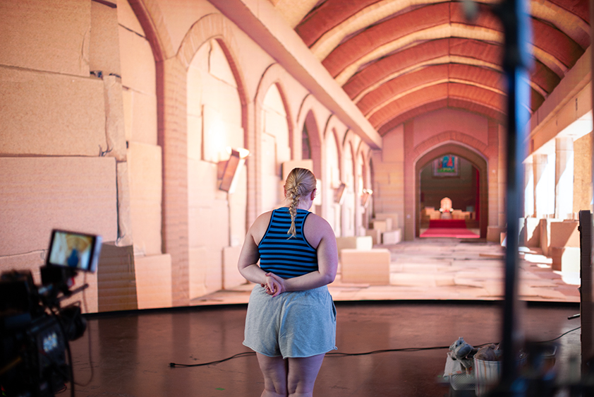 A college student stands on a virtual production stage. In front of her is a large, curved LED screen that is showing an elaborate castle scene.