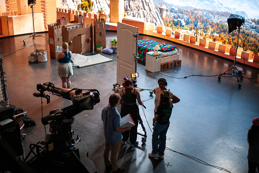 An elevated view of a college students working on a virtual production stage, including a large, curved LED screen showing a castle scene. A cardboard castle, a door and a child's bed and nightstand are on the stage.