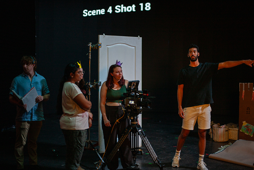 Four college students work with equipment on a virtual production stage, while an industry professional on the right gestures toward the stage. Text on the screen above them reads: Scene 4 Shot 18.