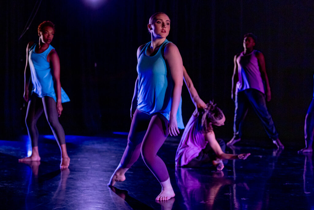 Four dancers are pictured performing on stage, with a blue light shining on them.