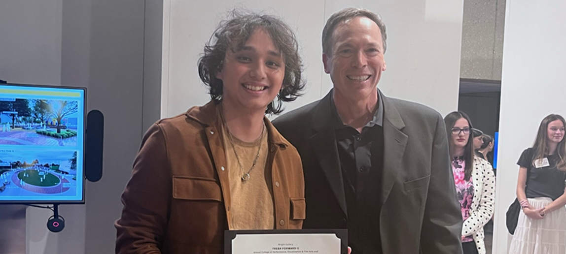 A college student holds a certificate for his award-winning project in an art exhibit. Standing next to him is the dean of the college.